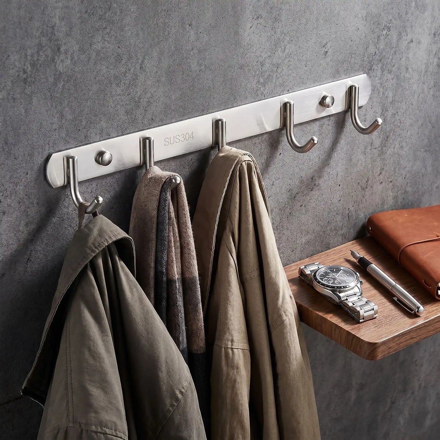 Détail du porte-manteau mural Le Pêcheur en acier inoxydable brossé argenté à côté d'accessoires élégants, illustrant la finition haut de gamme et la durabilité du matériau.