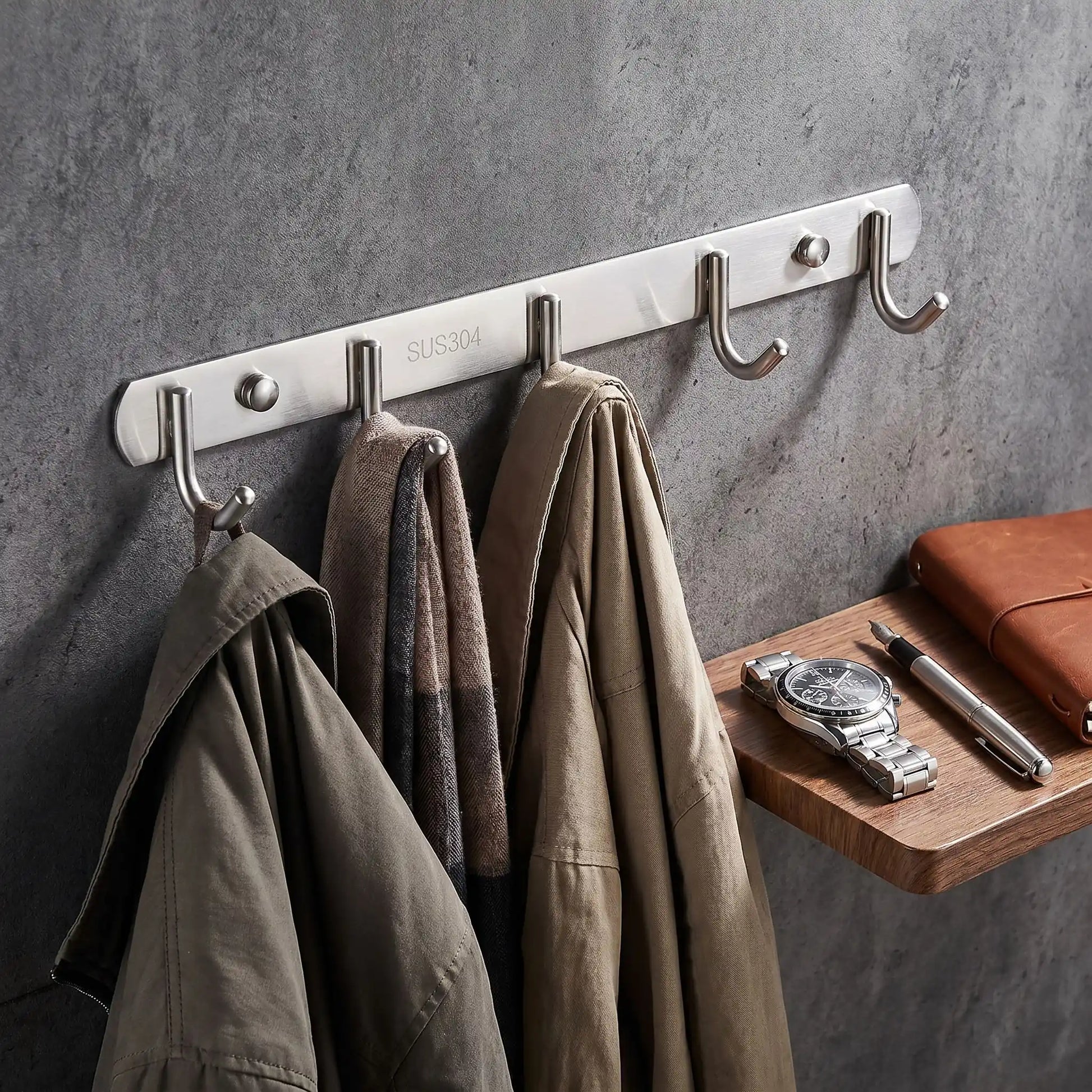 Détail du porte-manteau mural Le Pêcheur en acier inoxydable brossé argenté à côté d'accessoires élégants, illustrant la finition haut de gamme et la durabilité du matériau.