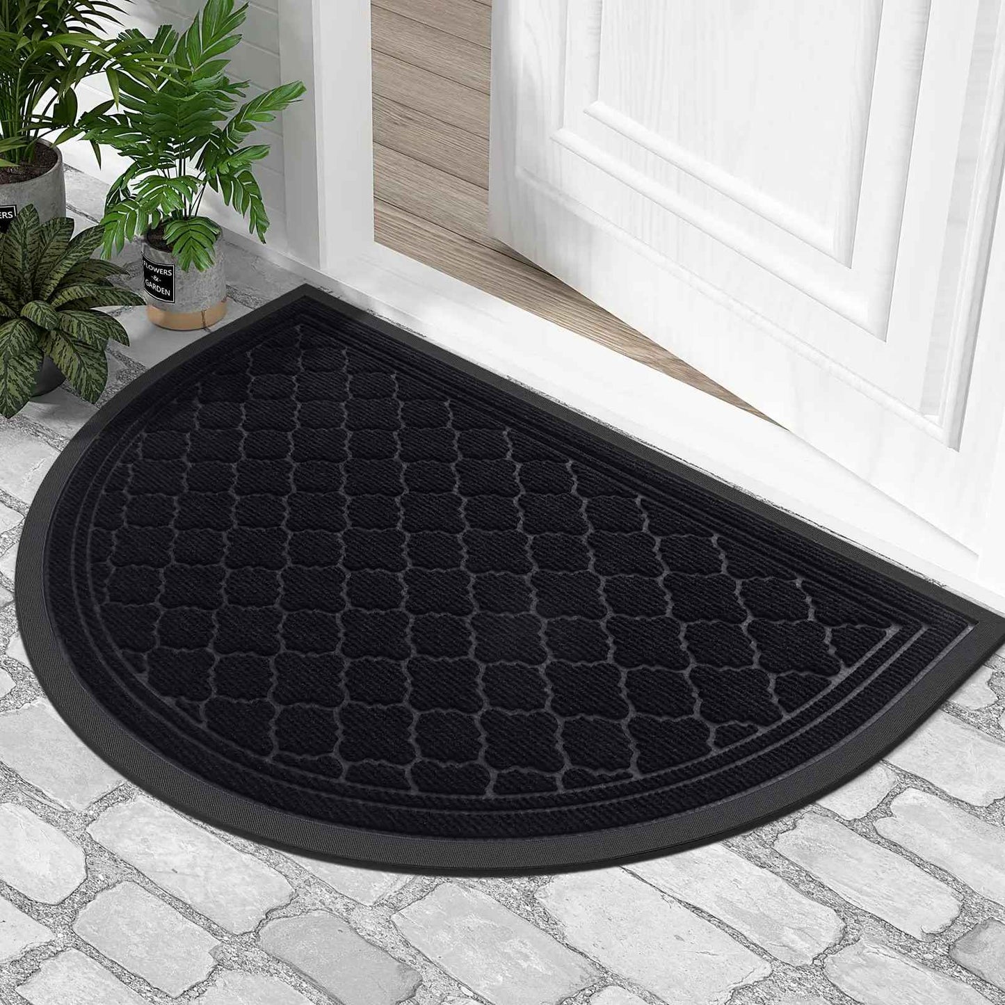 Tapis d'entrée demi-lune noir obsidienne avec bordure antidérapante posé sur un sol en pavés devant une porte blanche.