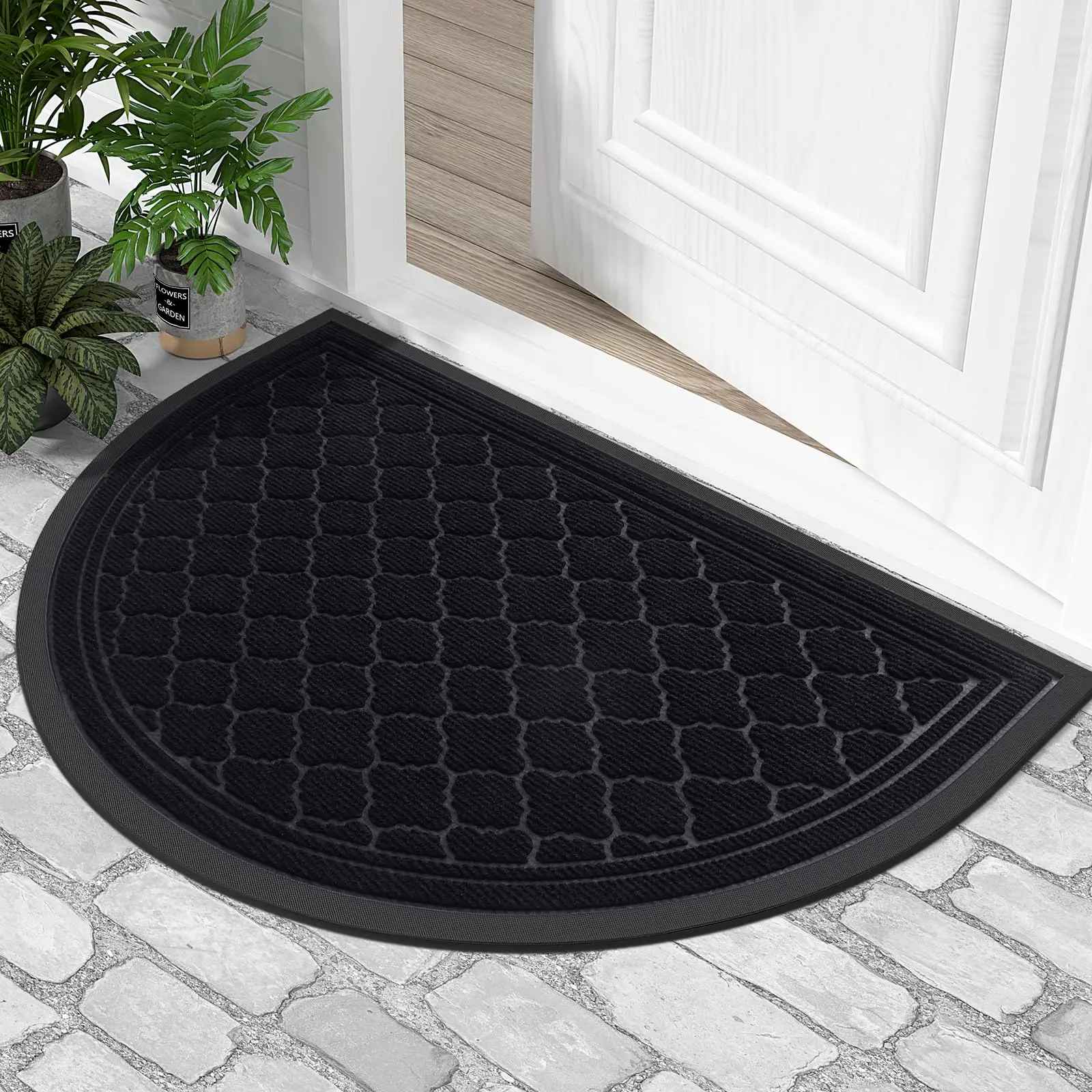 Tapis d'entrée demi-lune noir obsidienne avec bordure antidérapante posé sur un sol en pavés devant une porte blanche.